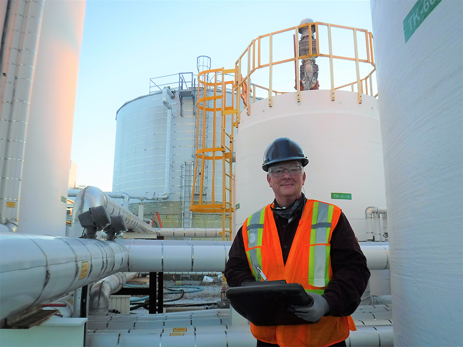 A man wearing a hard hat and reflective safety vest holding a clipboard. Behind him are building sized tanks connected by large pipes that snake into the foreground of the image