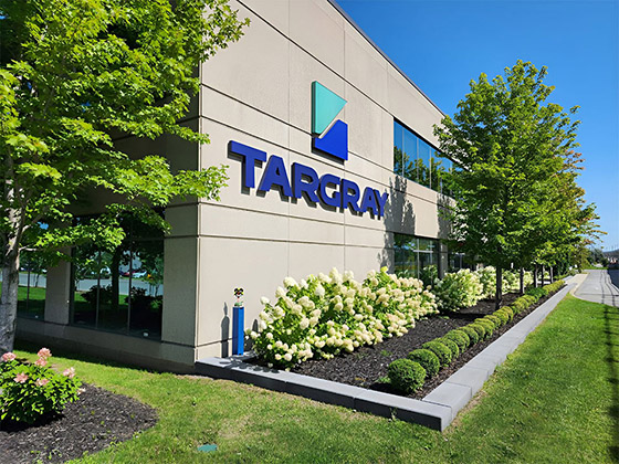 A large Targray logo mounted to the side of a nicely kept building in an office park. A few trees and a newly created garden line the building's wall.