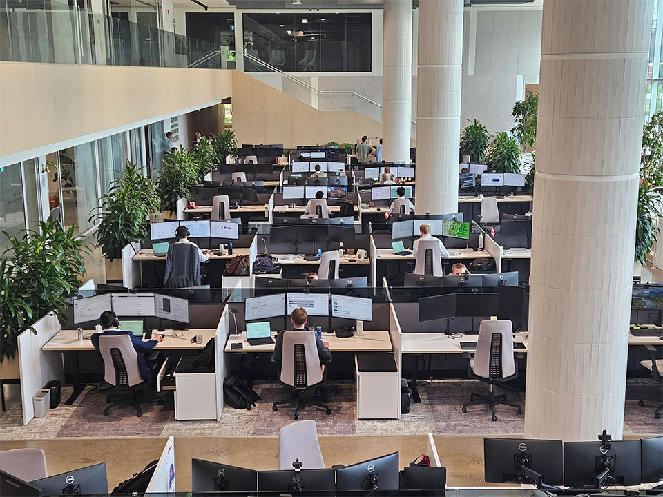 Overlooking a large naturally lit, open plan office. There are many plants among the rows of desks. There are a few emmployees working at clean workstations.